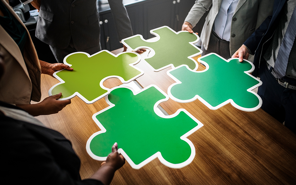 Business people assembling large green puzzle pieces on a wooden table, symbolizing teamwork and collaboration